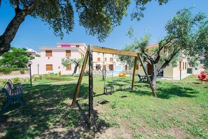 A playground with swings and a bench, surrounded by trees and buildings in the background. - Comfy Studio With Shared Pool (Orosei)