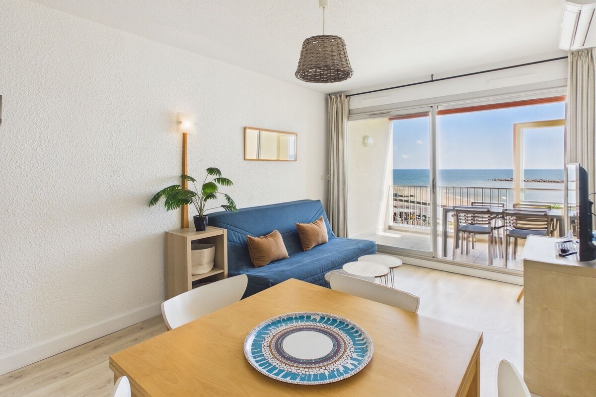 Un salon moderne avec un canapé bleu, une table à manger agrémentée d’un plateau décoratif, un luminaire suspendu et une vue sur la plage grâce à de grandes portes coulissantes.