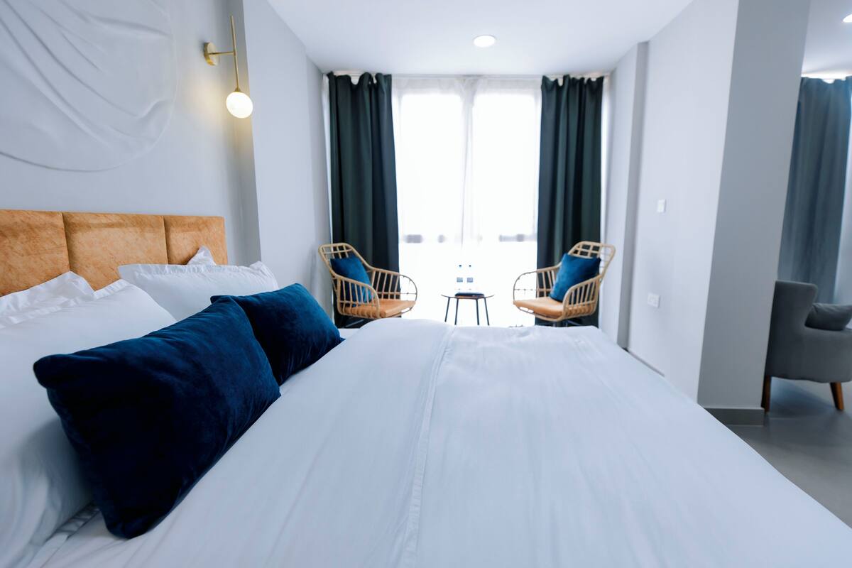A modern hotel room with a large bed, two wicker chairs, a small table, and a grey armchair.