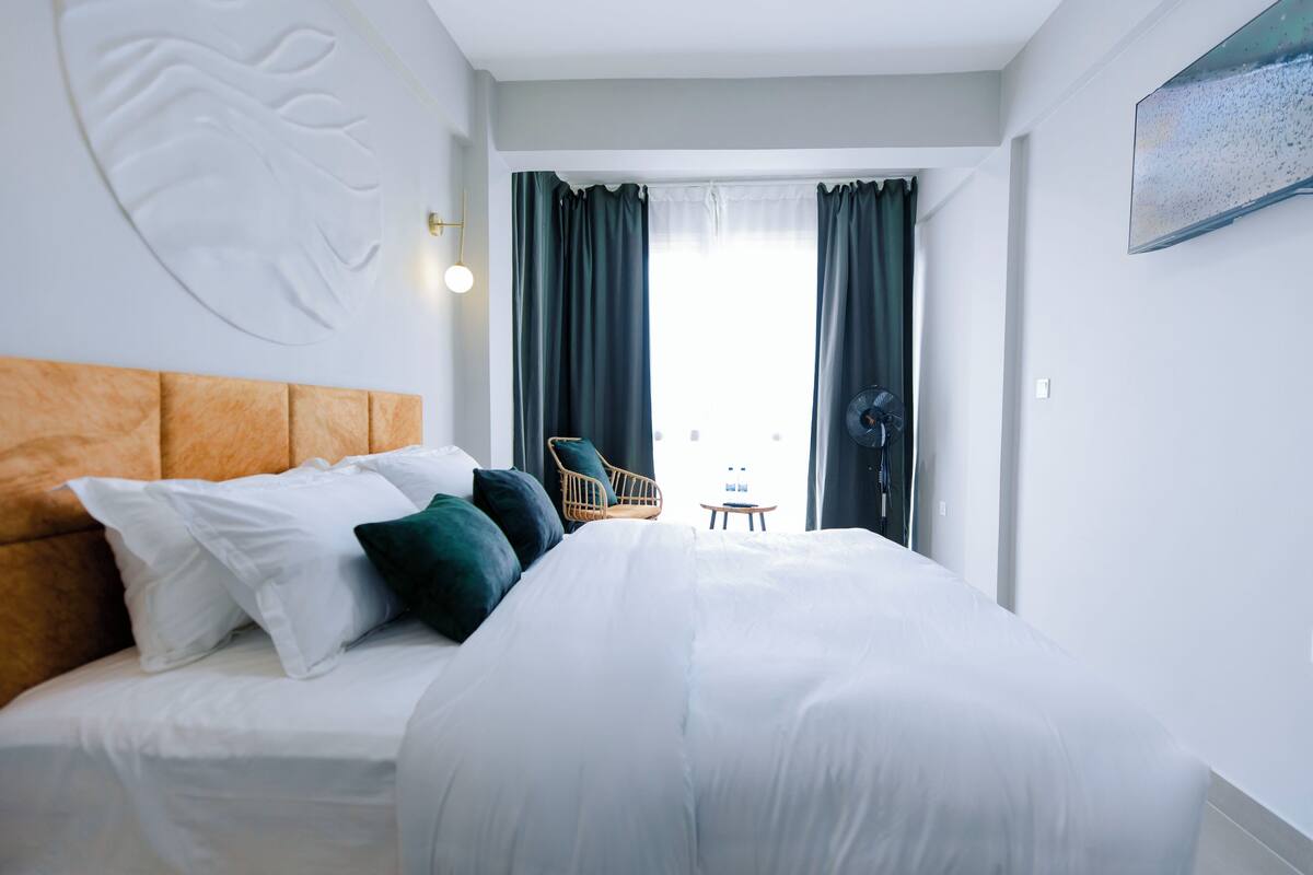 A hotel room with a large bed, a wooden headboard, a wicker chair, and a view of the city through the window.
