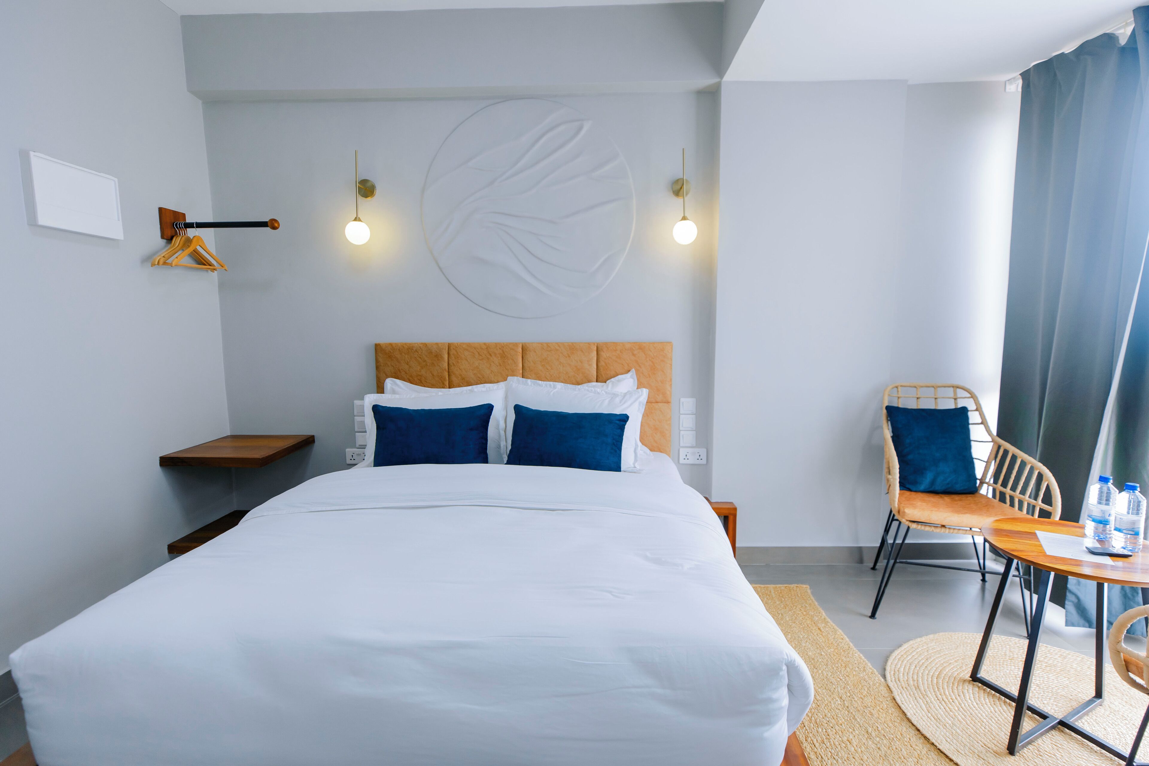 Une chambre d’hôtel bien rangée, avec un grand lit, une chaise, une petite table et un luminaire fixé au mur.