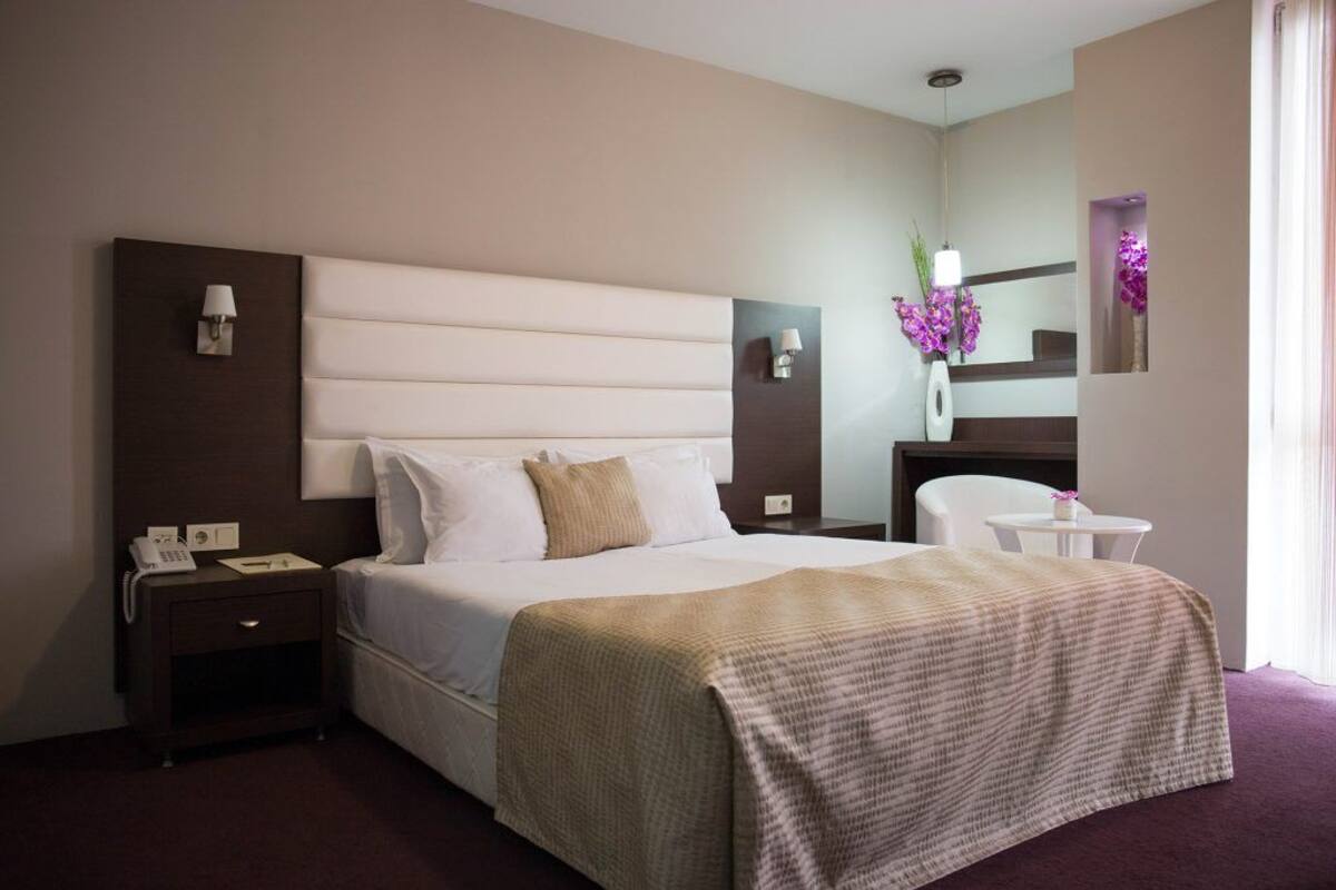 Une chambre d’hôtel avec un grand lit, une table de chevet, une commode, un miroir et une petite table sur laquelle est posé un vase de fleurs.
