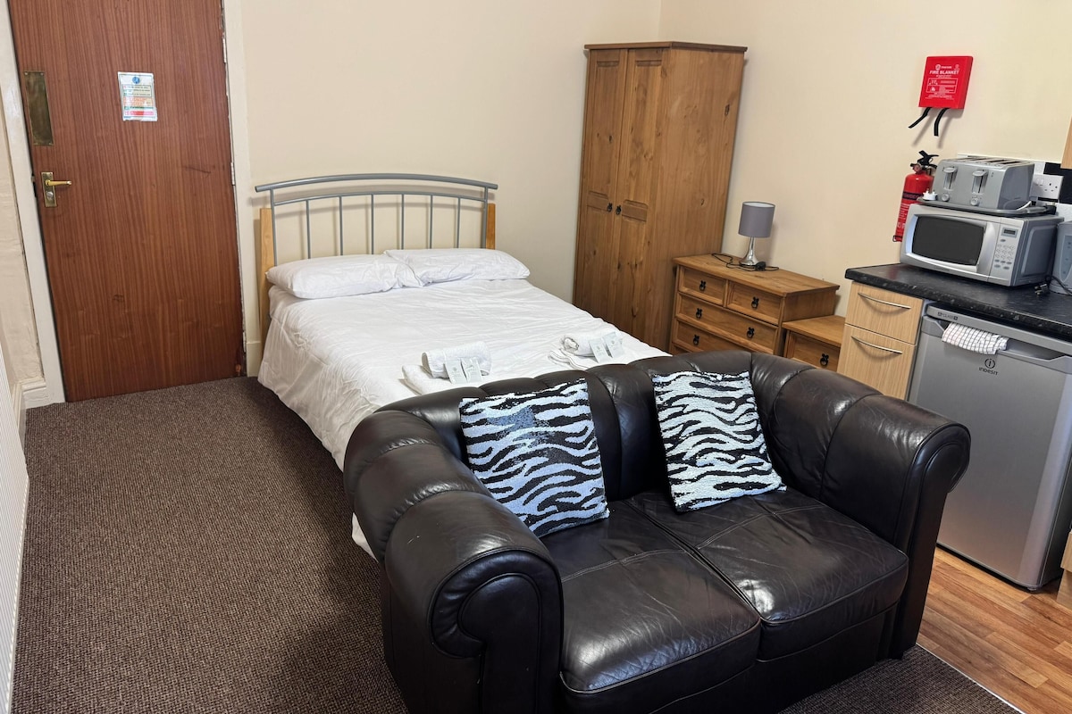A small, furnished room with a bed, a wooden wardrobe, a kitchenette with a microwave, and a fire blanket on the wall.