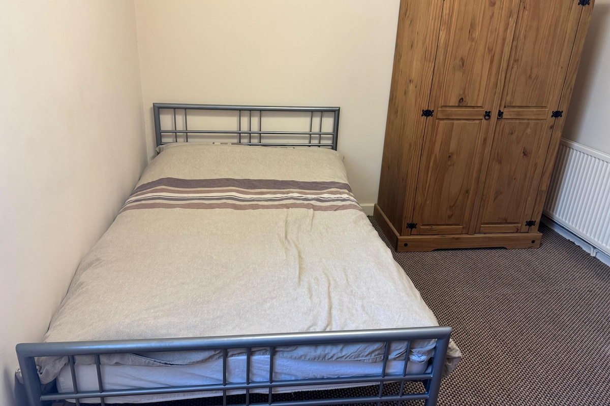 A single bed with a metal frame and a beige bedspread with brown stripes, a wooden wardrobe with black handles, and a carpeted floor.