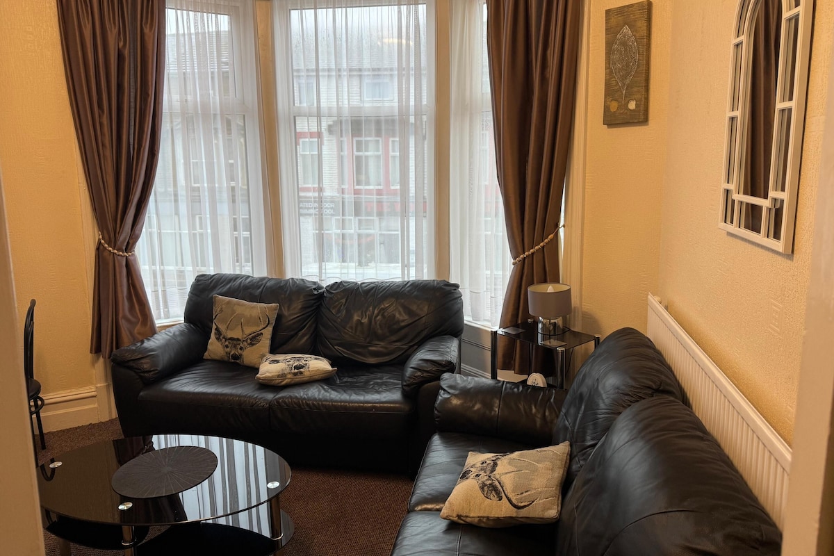 A living room with a black leather sofa, two matching cushions, a small round table, and a lamp on a side table.