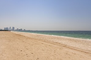 A wide sandy beach with a calm sea and a distant city skyline. - Luxe 4BR Villa Private Pool Beach Al Zorah (Ajman)