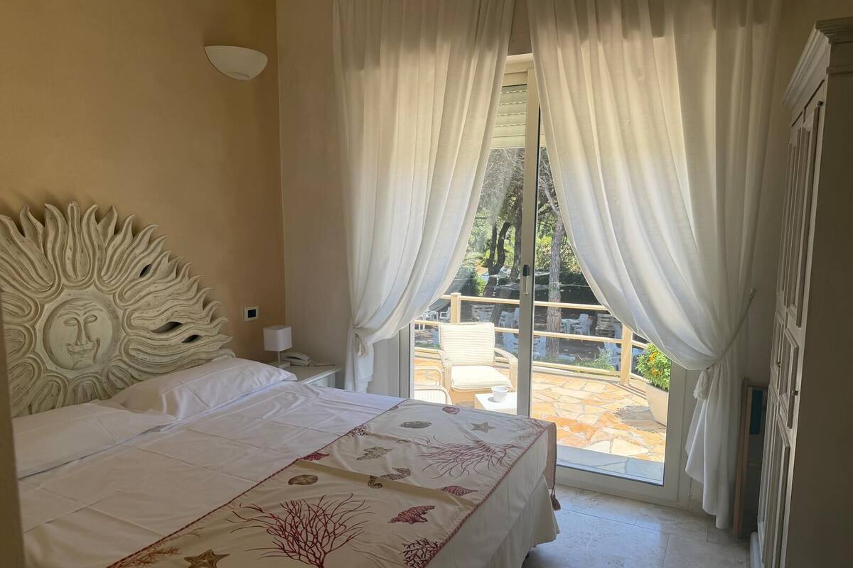 A bedroom with a large bed, a sunburst headboard, a balcony with outdoor furniture, and sheer curtains.