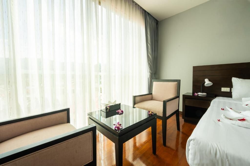 Une chambre d’hôtel avec un lit, une chaise, une petite table, une table de chevet et une fenêtre avec des rideaux légers.