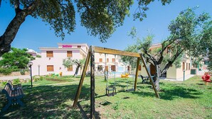 A playground with swings and a bench, surrounded by grass and trees, with buildings in the background. - Spacious Apartment With Shared Pool (Orosei)
