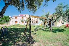 A playground with swings and a bench, surrounded by grass and trees, with buildings in the background. - Spacious Apartment With Shared Pool (Orosei)