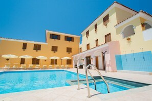 A swimming pool with sun loungers and umbrellas, surrounded by buildings with balconies and windows. - Spacious Apartment With Shared Pool (Orosei)