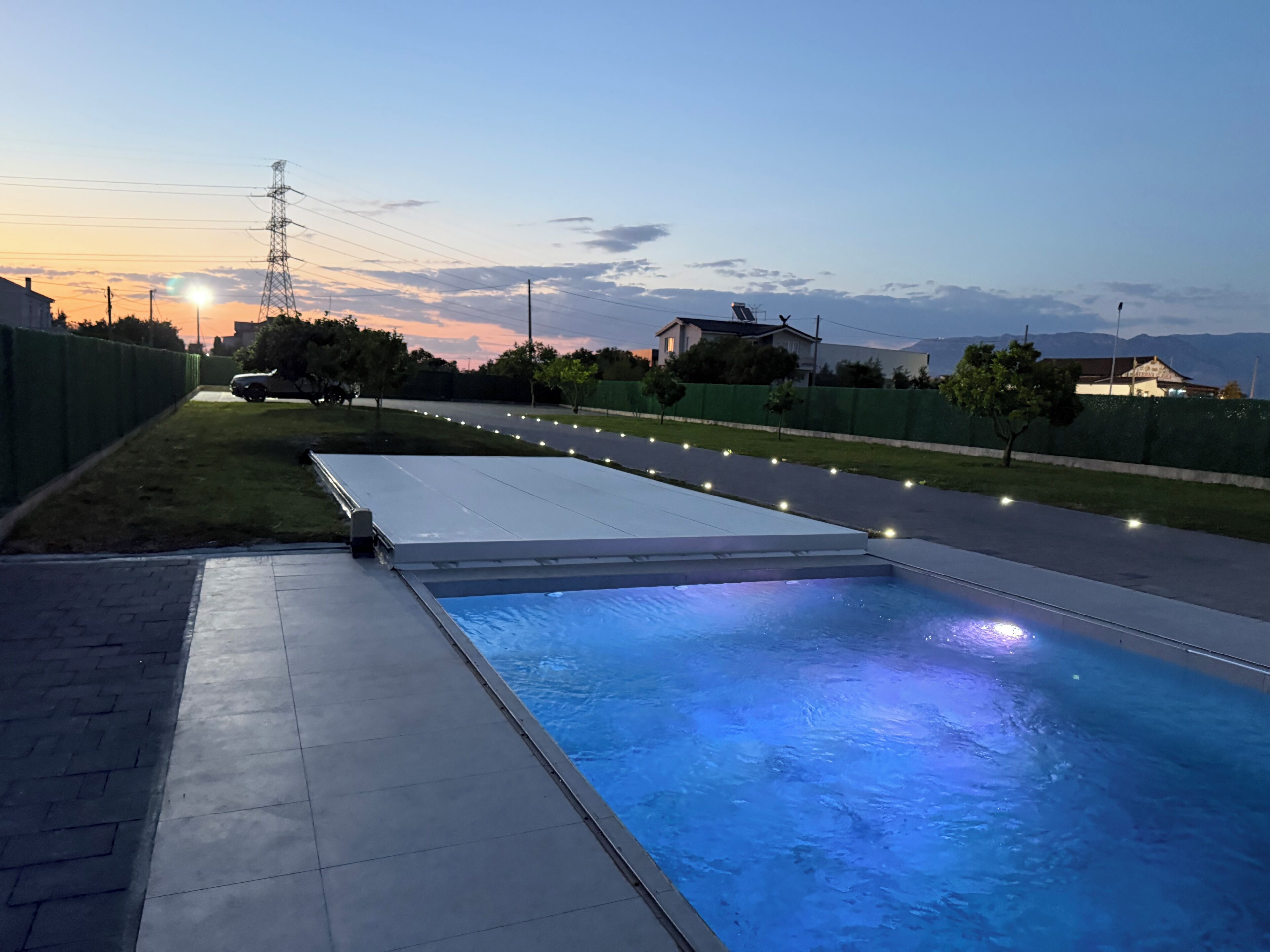 A swimming pool with a flat deck area, surrounded by a fence and a road with street lights.