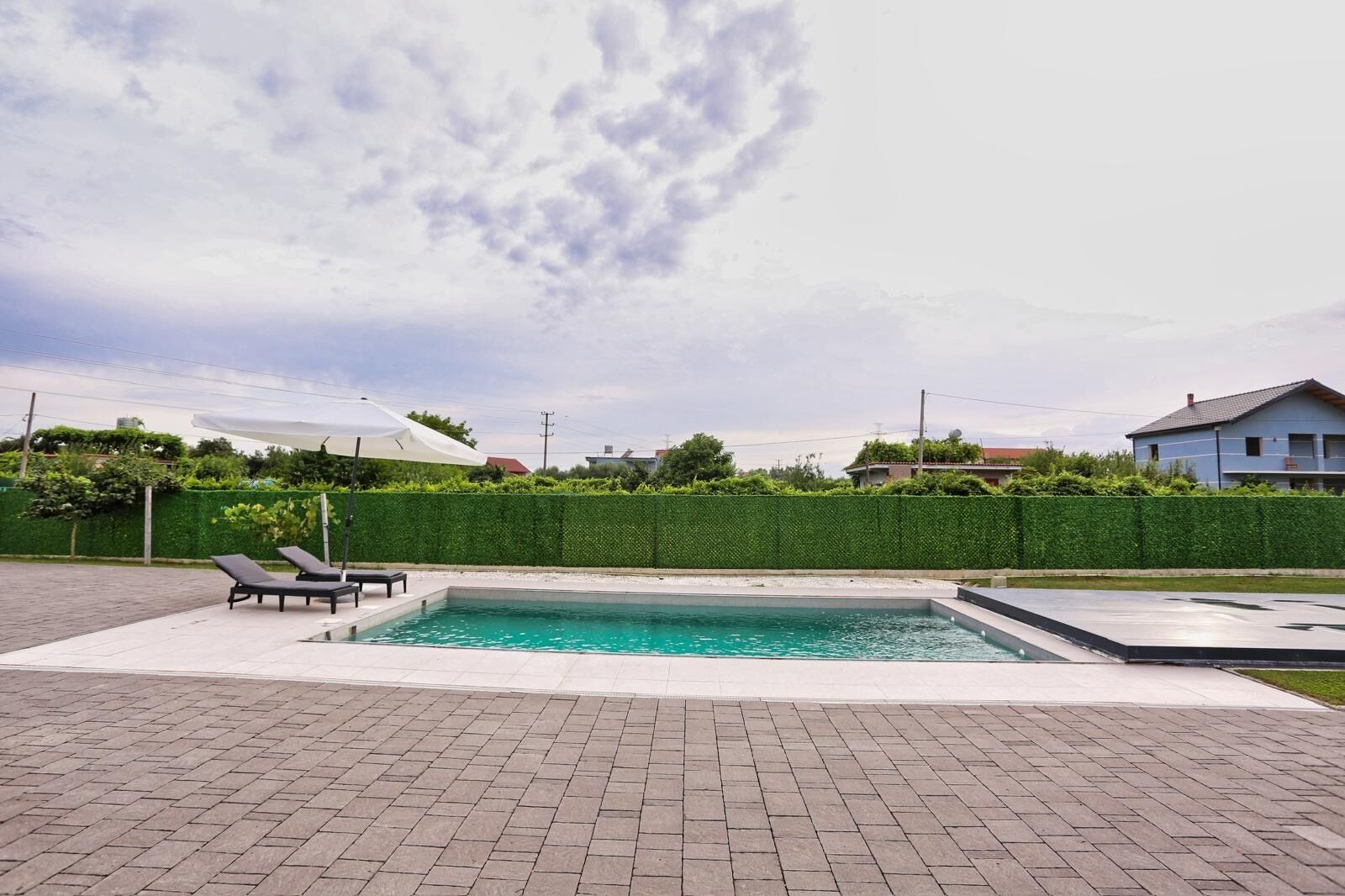 A rectangular swimming pool with a paved deck, two lounge chairs, a white umbrella, and a green hedge wall.
