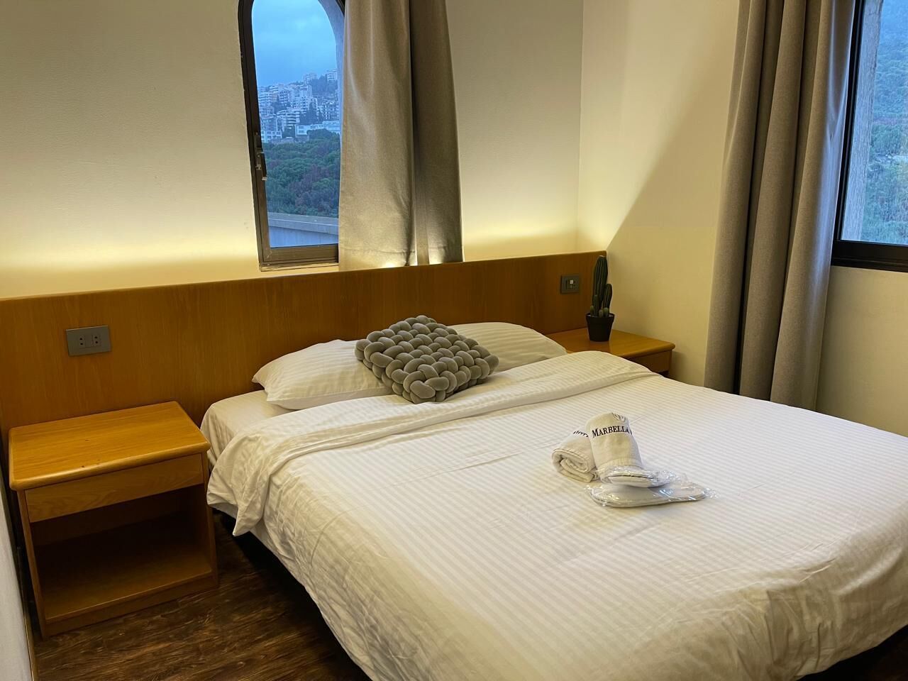 Ein Hotelzimmer mit Bett, Nachttisch, einem Fenster mit Vorhängen und Blick auf eine Stadtlandschaft.
