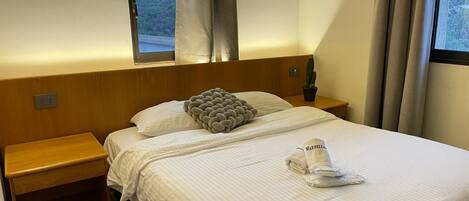 Habitación de hotel con cama, mesita de noche, ventana con cortinas y vista a la ciudad.