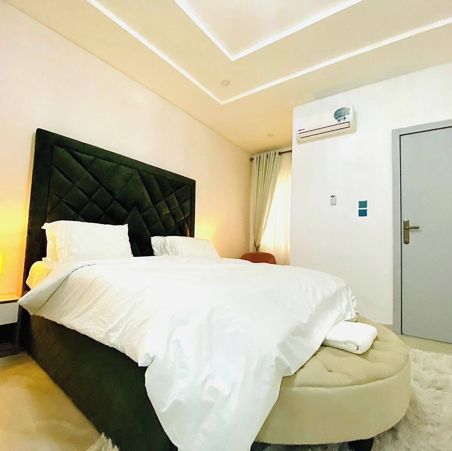A modern bedroom with a large bed, a dark headboard, a bedside table, an air conditioning unit, and a door.