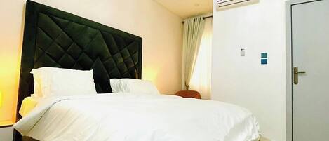 A modern bedroom with a large bed, a dark headboard, a bedside table, an air conditioning unit, and a door.