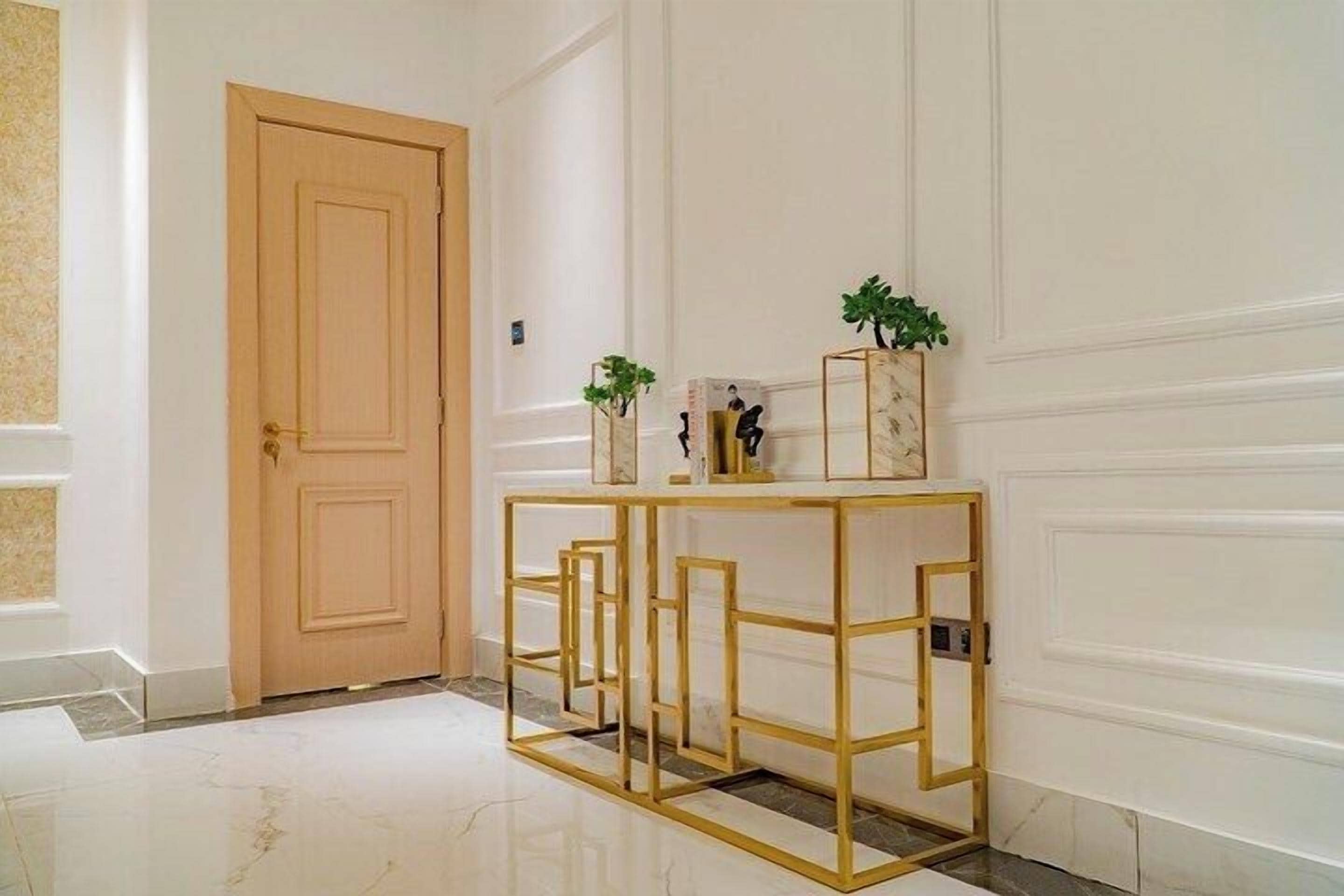Un couloir avec une porte en bois, un sol en marbre et une console aux motifs décoratifs, dont le cadre est doré.