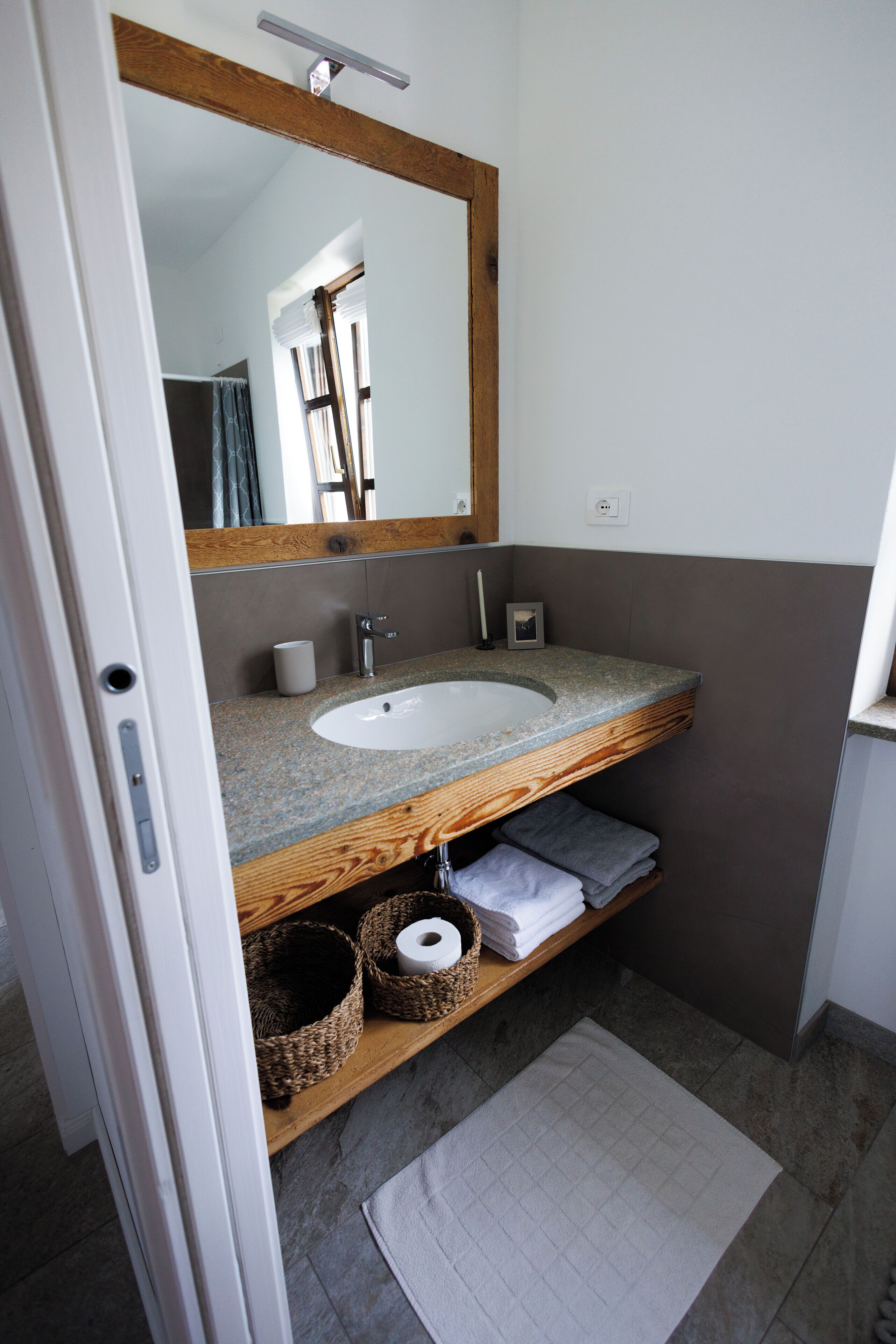 A bathroom with a wooden vanity, a white sink, and a mirror. There are baskets under the vanity and a towel on the floor.