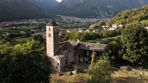 Un edificio de piedra histórico con una torre, rodeado de abundante vegetación y montañas al fondo.