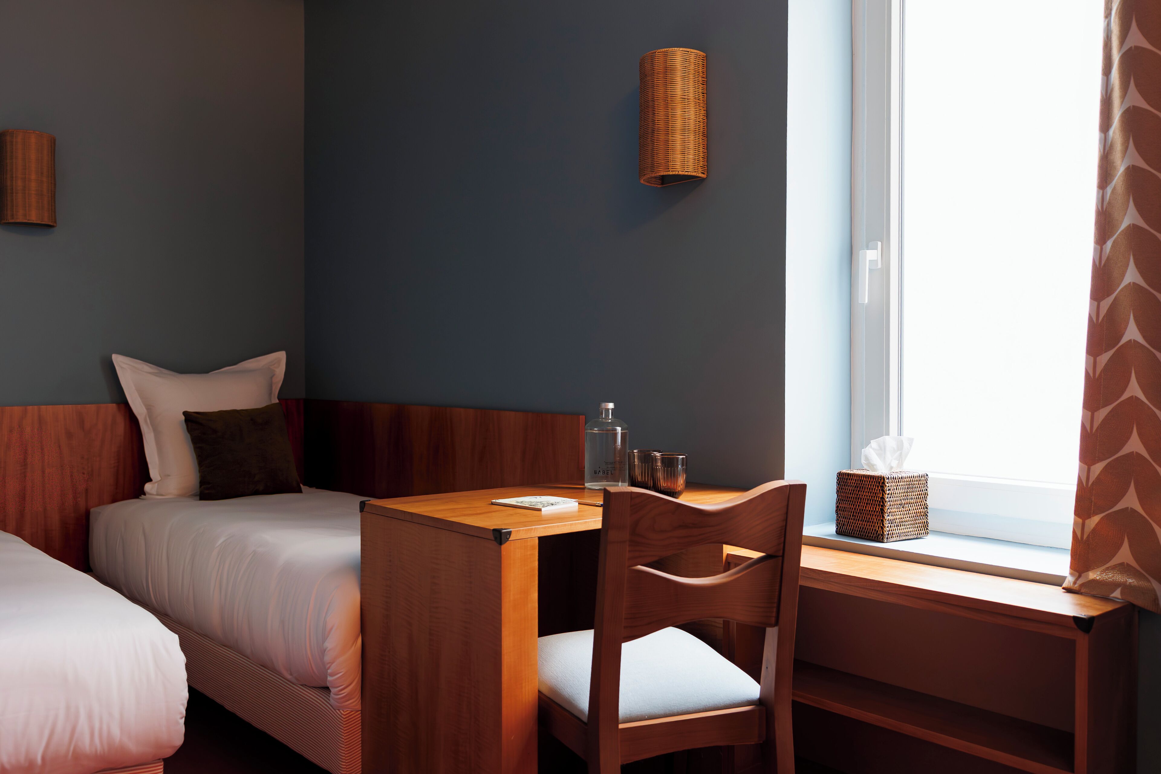 Une chambre d’hôtel avec deux lits, un bureau en bois, une chaise, une lampe et une fenêtre avec des rideaux.