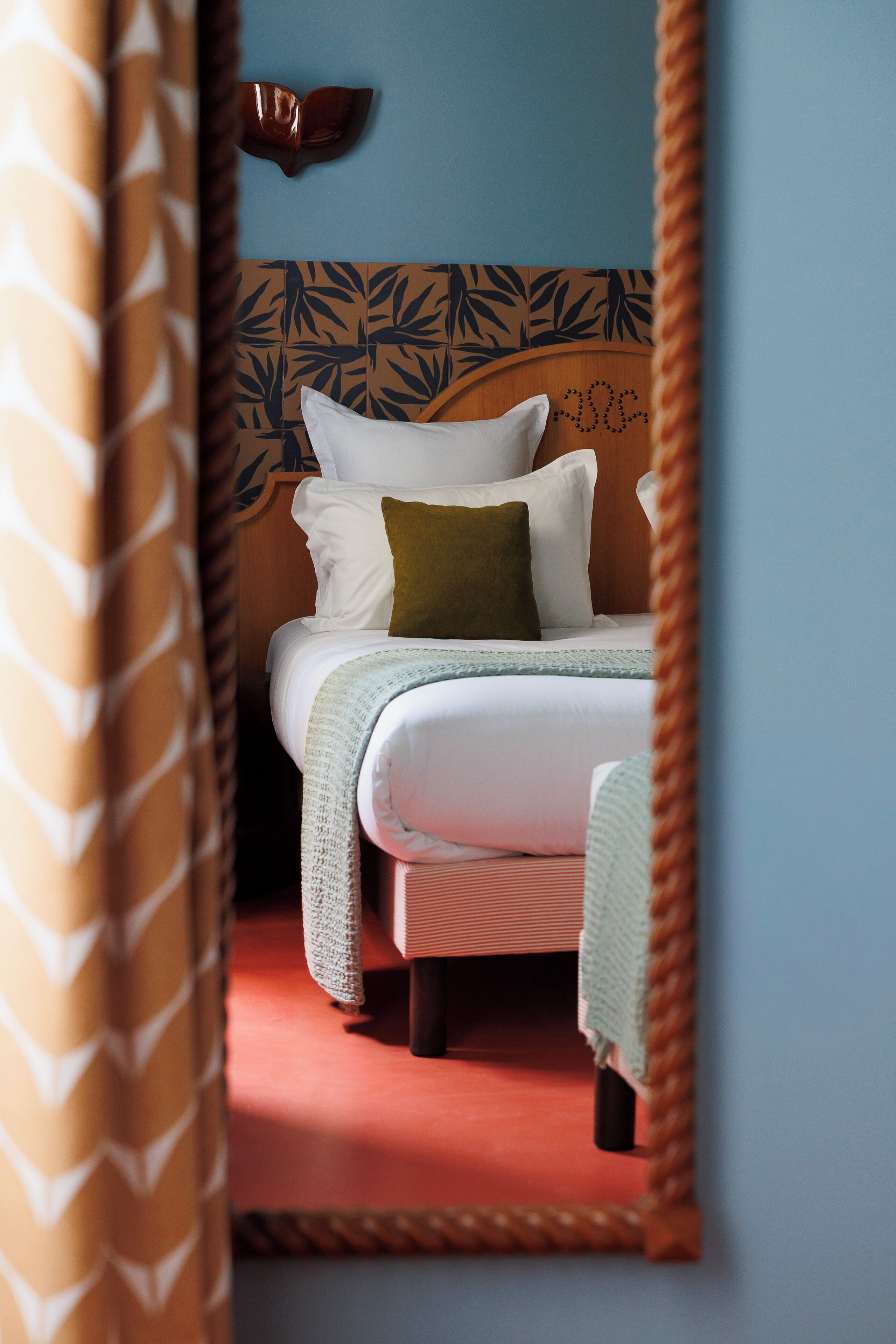 A bedroom with a bed, pillows, a headboard with a leaf pattern, and a mirror reflecting the room.