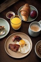 Un desayuno con croissants, fruta, un vaso de zumo de naranja y un plato con bacon y un huevo.