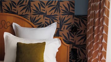 Een net opgemaakt bed met witte en groene kussens, een houten hoofdbord met een bladmotief, een houten nachtkastje en een gordijn met een patroon.