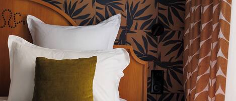A neatly made bed with white and green pillows, a wooden headboard with a leaf pattern, a wooden side table, and a patterned curtain.