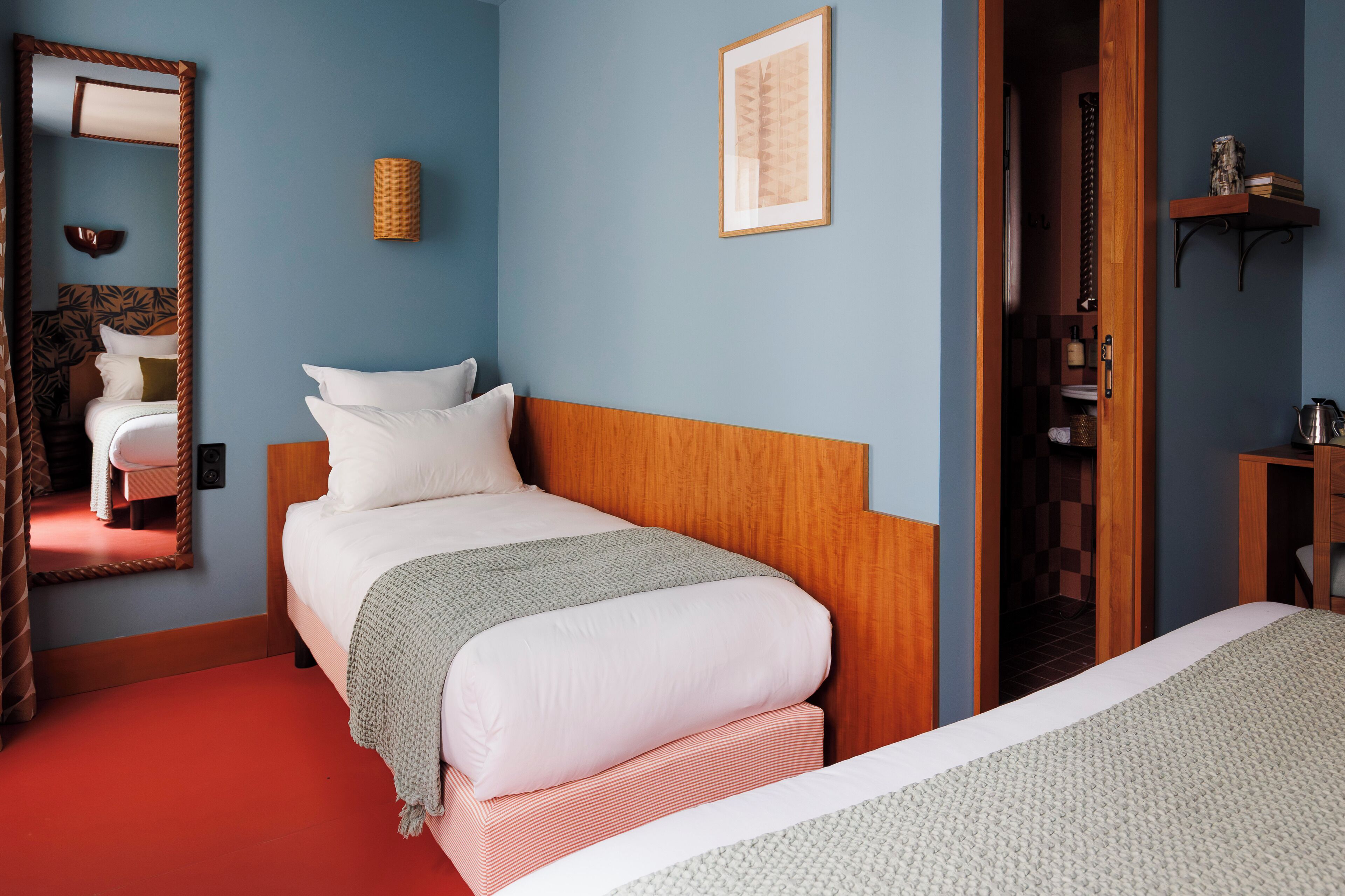 A hotel room with a large mirror, a bed with white and red bedding, a wooden headboard, and a small shelf with books and a coffee pot.