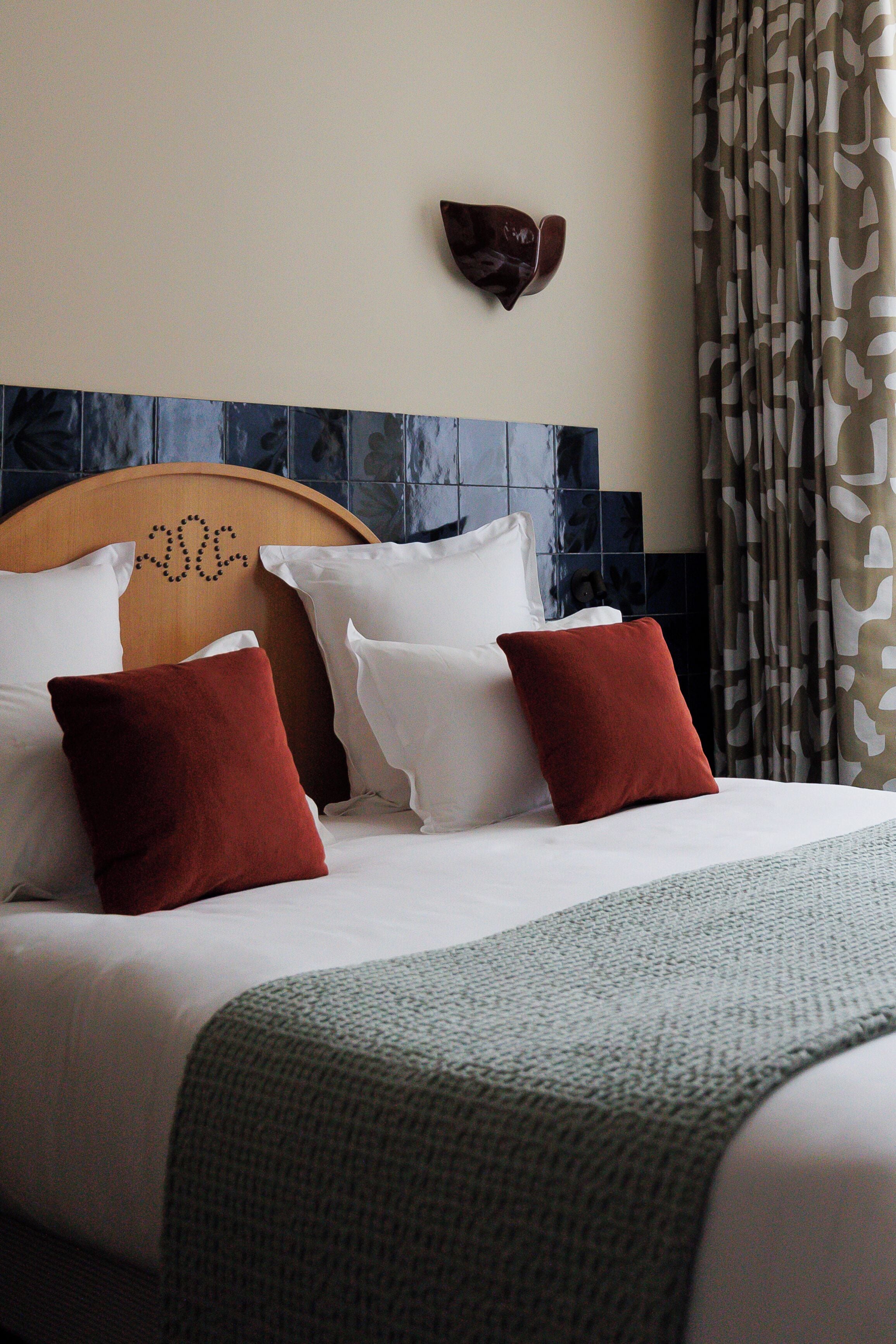 Una cama bien hecha con almohadas blancas y rojas, un cabecero de madera y una cortina estampada.