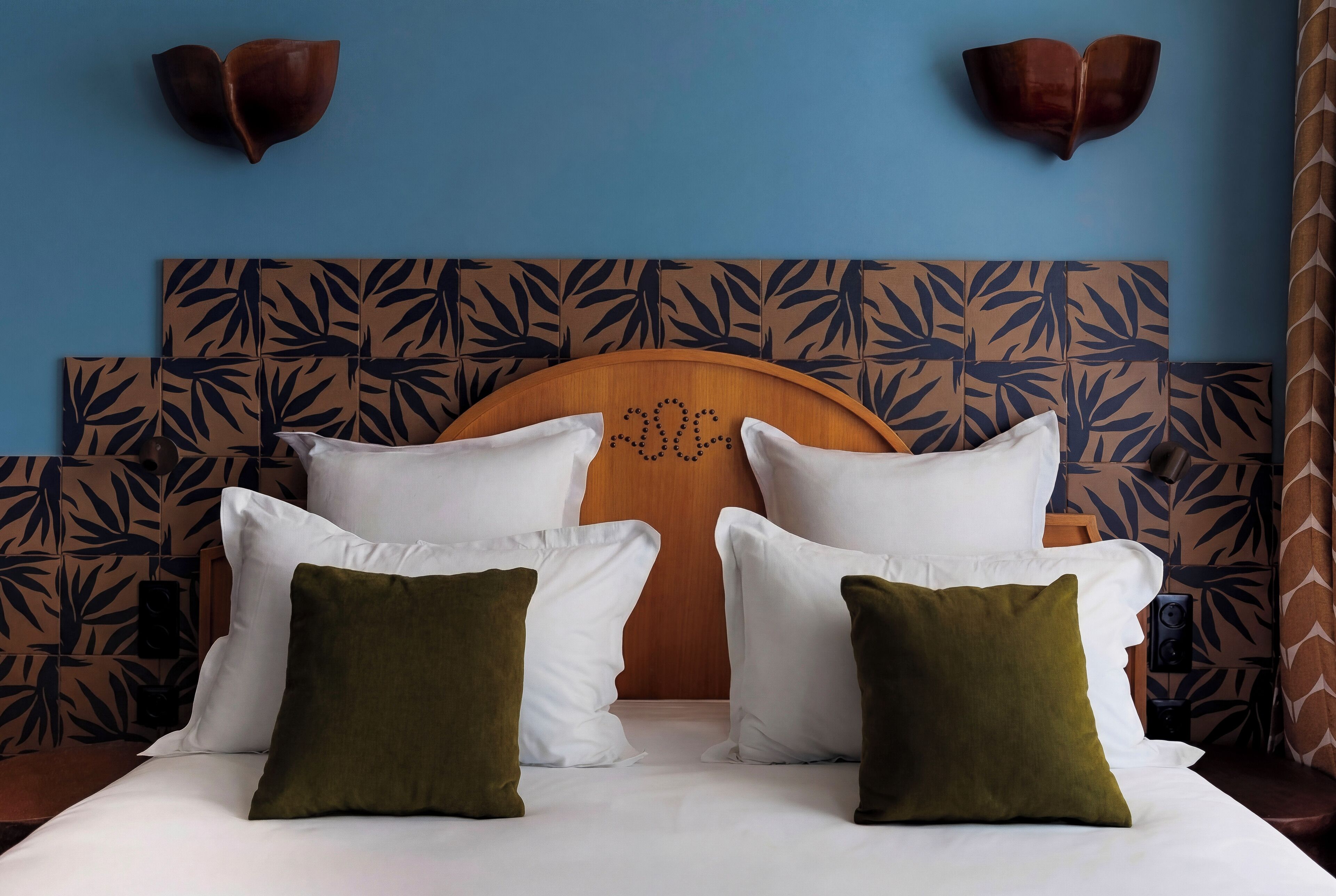 Una cama con almohadas blancas y verdes, cabecera de madera con diseño de hojas y lámparas fijadas a la pared.