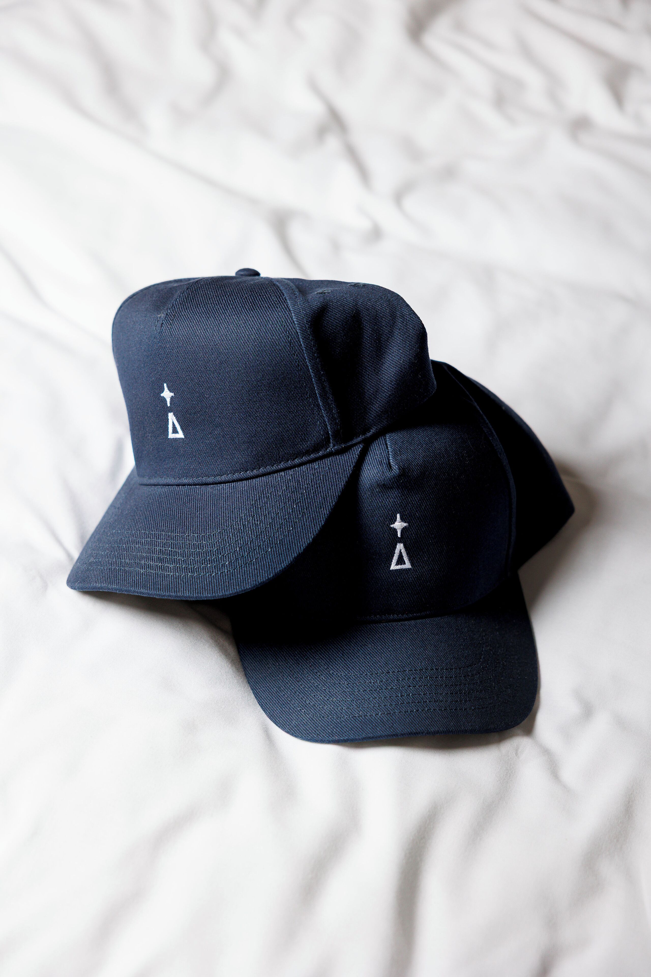 Dos gorras de béisbol azul marino con estrellas y triángulos bordados en blanco sobre una superficie de tela blanca.