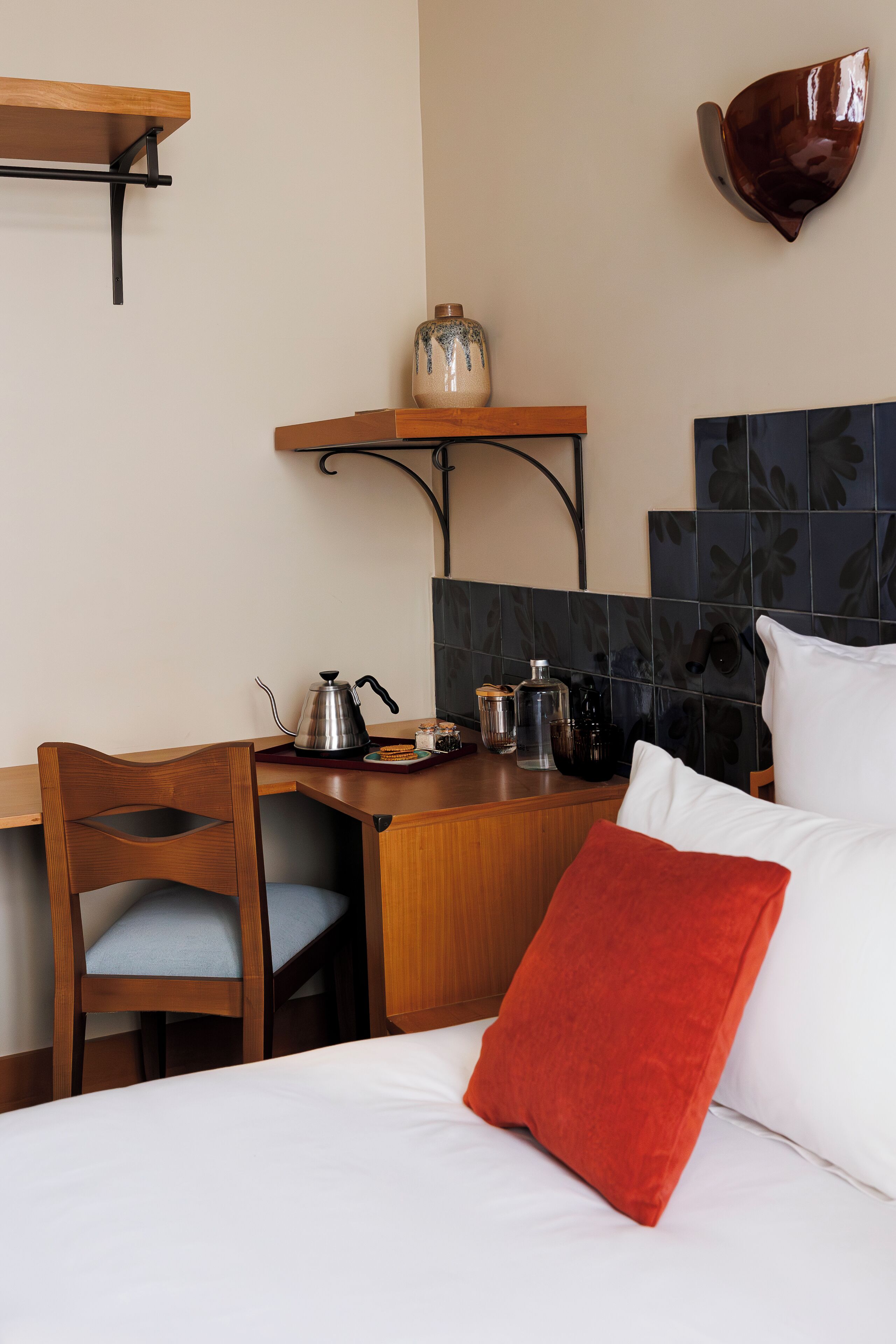 A hotel room with a wooden desk, a chair, a bed with white bedding and a red pillow, a coffee pot, a kettle, a jar, and a wall-mounted bowl.