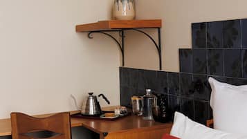 A hotel room with a wooden desk, a chair, a bed with white bedding and a red pillow, a coffee pot, a kettle, a jar, and a wall-mounted bowl.