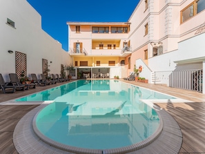 Una piscina con sillones de descanso y un área de estar cubierta, rodeada por un edificio con balcones y fachada blanca.