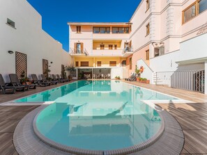 A swimming pool with lounge chairs and a covered seating area, surrounded by buildings with balconies and a clear blue sky. - Comfy 4 Guest Apartment With Pool (Santa Teresa di Gallura)
