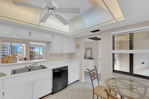 A modern kitchen with white cabinetry, a dishwasher, a ceiling fan, and a dining area with a glass table and chairs. - SANDARAC 604b BEACHFRONT 2B/2B VACATION CONDO w/  GULF VIEW, RESORT POOL! (Fort Myers Beach)