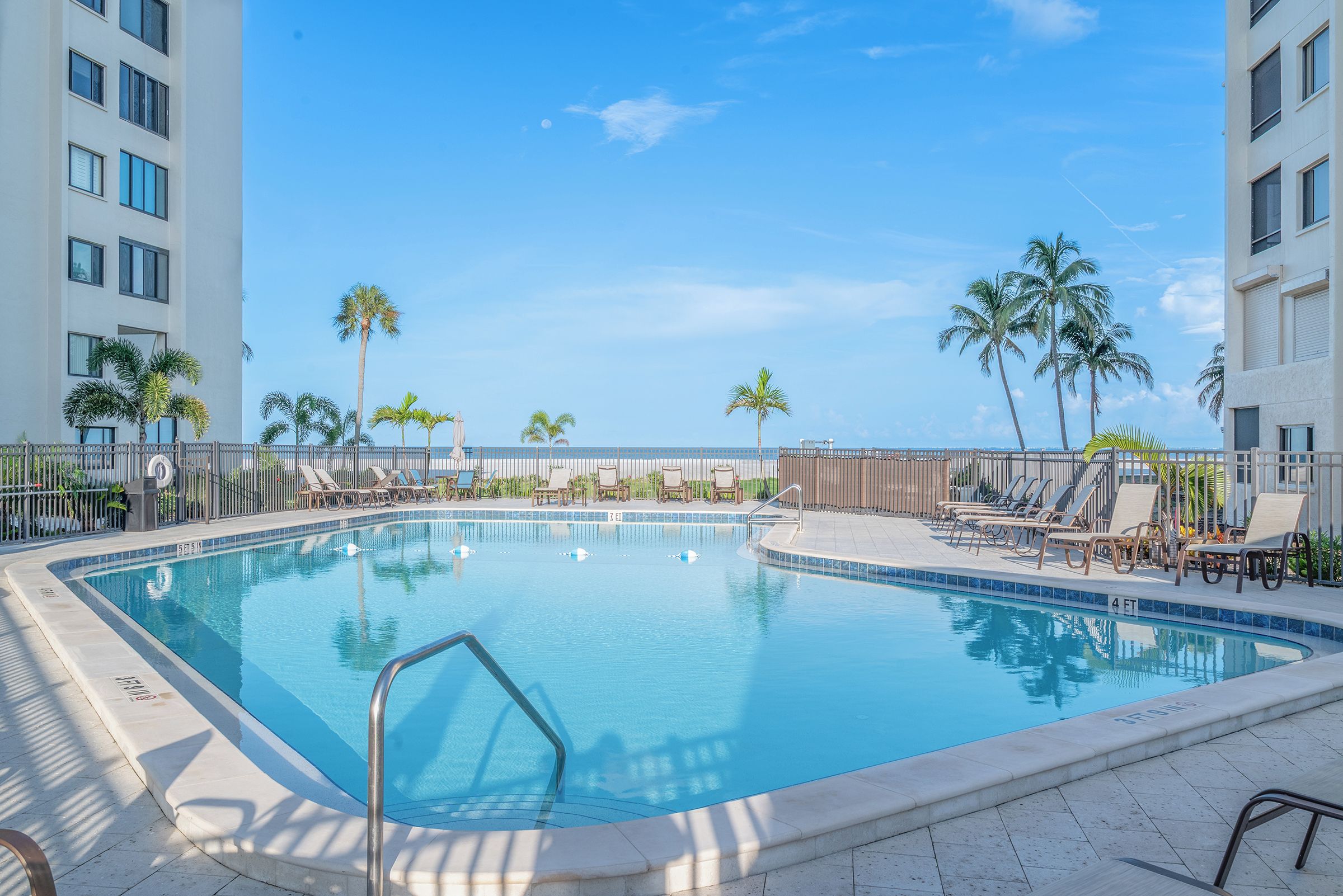 A rectangular swimming pool with a metal ladder, surrounded by lounge chairs and palm trees, with a clear sky in the background.