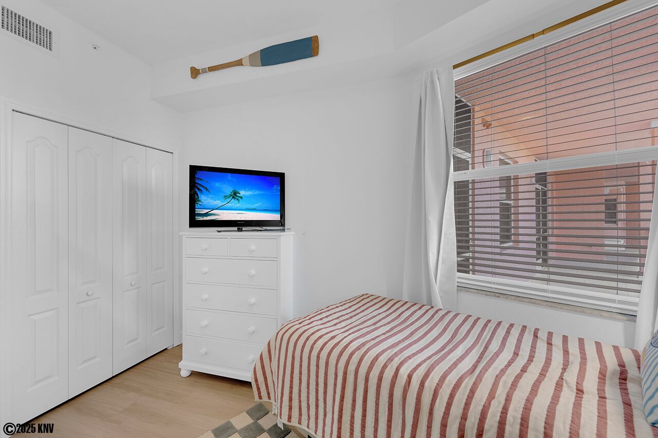Ein Schlafzimmer mit Fernseher auf einer Kommode, einem Bett mit gestreifter Bettwäsche, einem Fenster mit Jalousien und einem weißen Kleiderschrank.