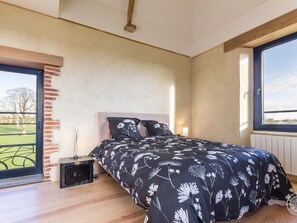 A bedroom with a large bed, a nightstand, a window with a view of a green landscape, and a brick wall. - Historic house in Normandy with panoramic views (Belval)