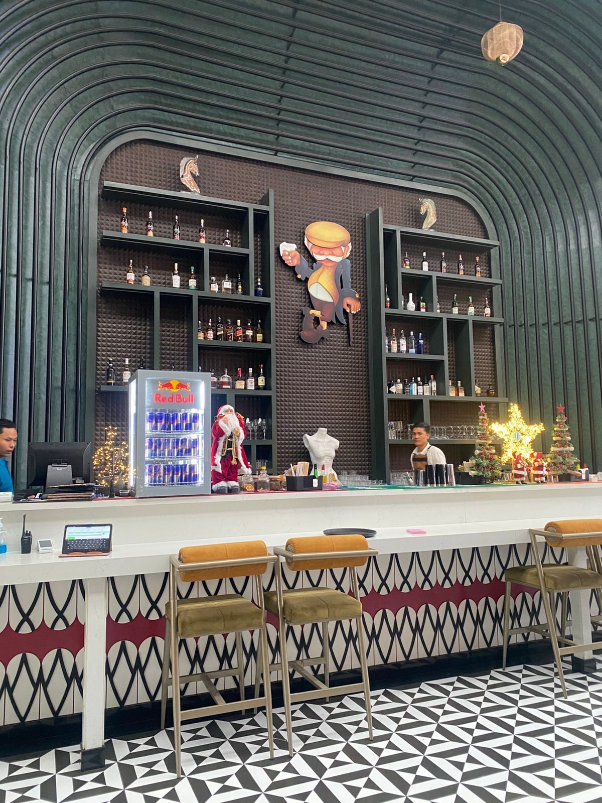 A bar area with shelves of bottles, a Red Bull refrigerator, and a Santa Claus figure.