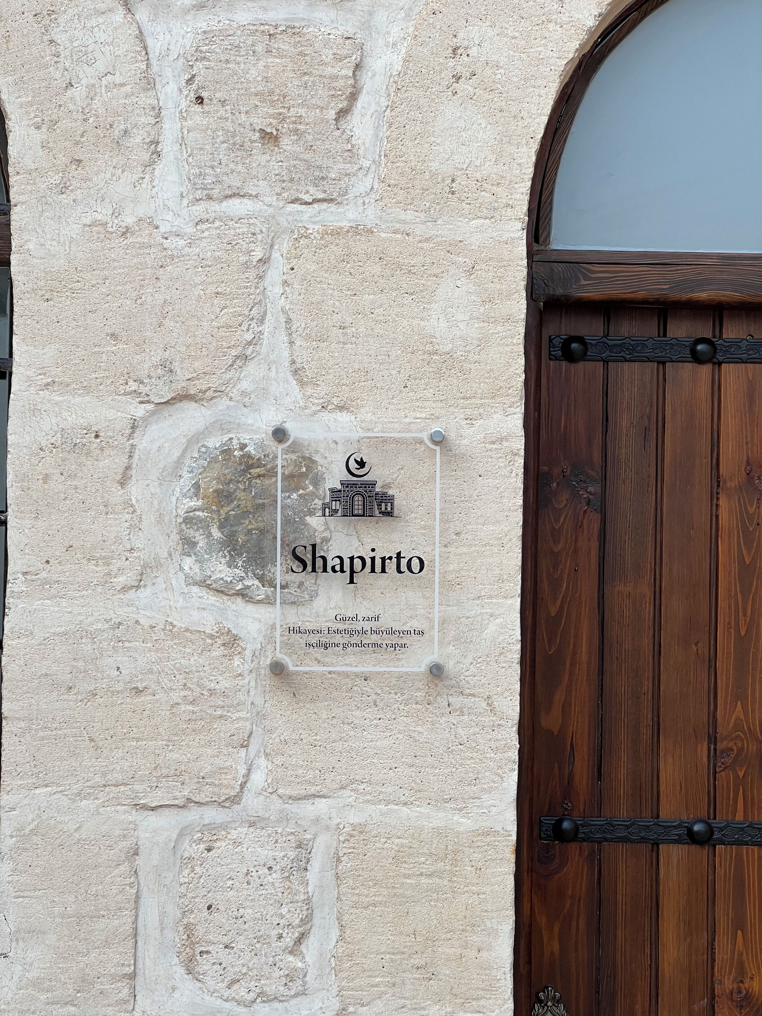 Un muro de piedra con una puerta de madera y un letrero que dice "Shapirto" con una descripción en turco.