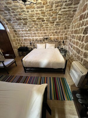 Une chambre avec un mur en pierre, un lit avec une tête de lit, une table de chevet, une chaise, un tapis à rayures et un appareil de climatisation.
