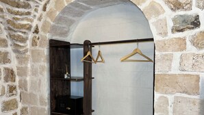 Ein Zimmer mit Steinwänden, einer hölzernen Sitzbank, einem Kleiderständer mit Kleiderbügeln und einem dunklen Holzschrank.