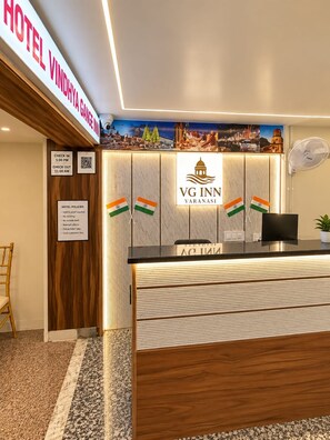 A hotel reception desk with a check-in/check-out sign, a digital display, and a wall with a scenic poster. - Hotel Vindhya Ganga Inn (Varanasi)