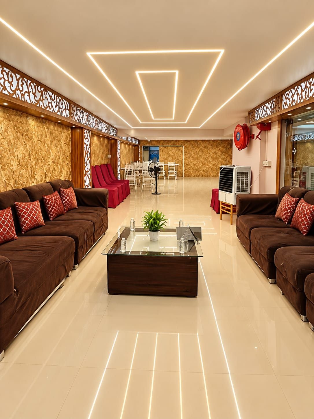 Un vestíbulo moderno con una mesa de centro de cristal, zonas de estar con sofás marrones y cojines rojos, y un diseño de pared con estampado.