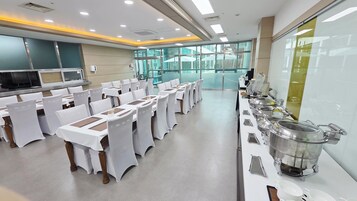 Un salón de banquetes con filas de mesas dispuestas para un evento formal, equipado con una barra de buffet en un lateral.