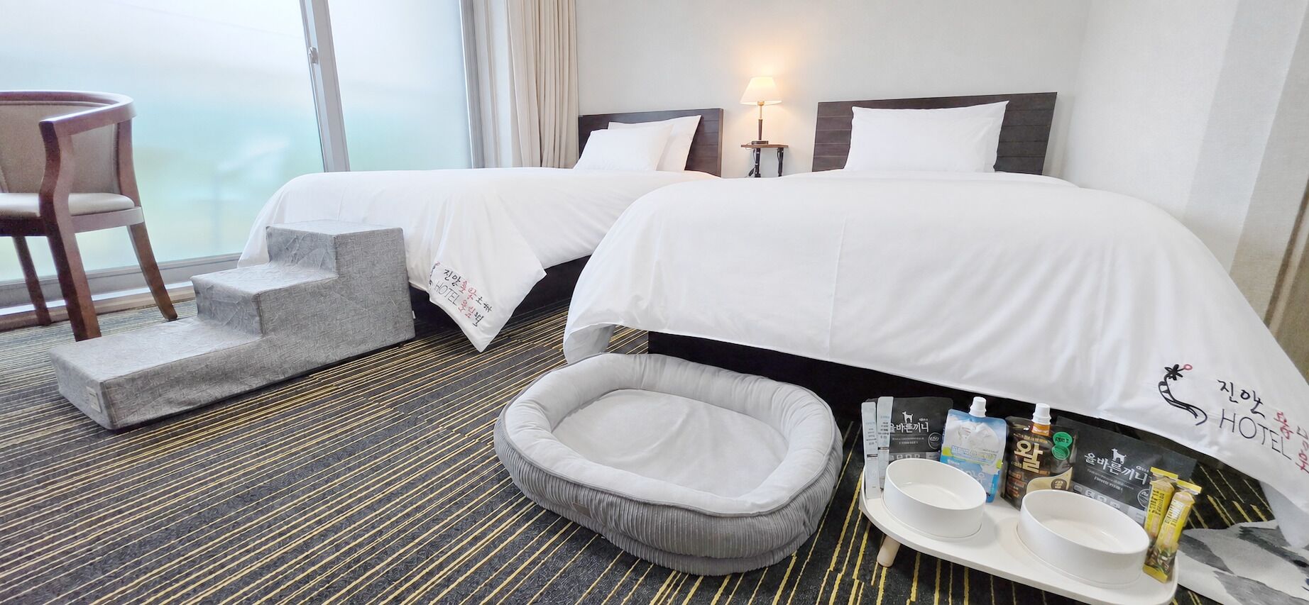 A hotel room with two beds, a pet bed, a small table with toiletries, and a chair.
