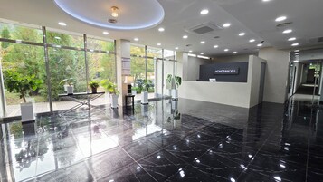 Un área de recepción moderna con suelo de mármol negro, amplias ventanas que ofrecen vistas a zonas verdes y un mostrador de recepción con el rótulo "HONGSAM VILL".